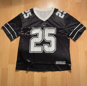 Svart fotbollströja Shein Teen 13Y - Oanvänd svart fotbollströja från Shein med stora vita siffror 25 på bröstet, ryggen och axlarna. Tröjan har vita detaljer, V-ringning och korta ärmar med dubbla ränder.