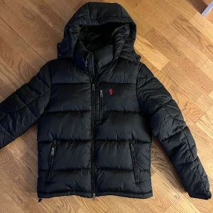 Svart pufferjacka från Polo Ralph Lauren - Svart pufferjacka från Polo Ralph Lauren med röd broderad logga på bröstet. Perfekt skick. Jackan har huva, flera dragkedjefickor och quiltad design. Storlek S.