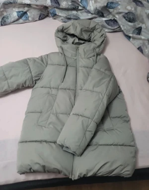 Ljusgrå pufferjacka från Amisu - Säljer en ljusgrå pufferjacka från Amisu i storlek 34. Jackan har huva med dragsko, långa ärmar och dragkedja framtill. Den är quiltad och har två fickor på sidorna. Perfekt för kalla dagar när du vill hålla dig varm och snygg. Använt bara 1-2 gånger och rätt så varm. inga täcken på användning, som ny.
