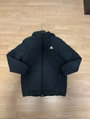Svart pufferjacka från Adidas - Svart pufferjacka från Adidas med huva och dragkedja framtill. Jackan har quiltad design, två sidofickor och Adidas-logga på bröstet. Perfekt för kalla dagar och riktigt skön att bära. Materialet är polyester och jackan har lång ärm.