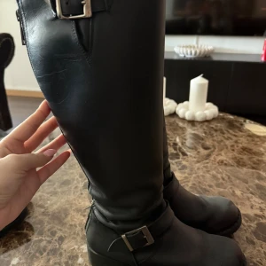 Svarta höga stövlar från Johnny Bulls - Snygga svarta stövlar från Johnny Bulls med högt skaft och coola metallspännen vid vrist och skaft. Gjorda i slätt skinn som ger en stilren look. Perfekta för dig som gillar klassisk och edgy stil. Rund tå och platt sula för bekväm användning.