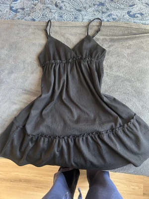 Svart kort klänning med volang - Supersöt svart kort klänning med tunna axelband och v-ringad hals. Klänningen har en volangkant nertill och lätt rynkade detaljer vid midjan och kjolen. Perfekt för varma dagar och enkel att styla med sneakers eller sandaler. Den har ingen lapp men sitter bra på mig som bär xs-s 