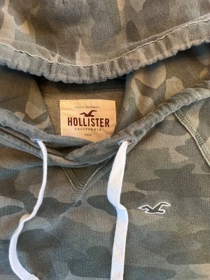 Camo hoodie av Hollister - Snygg hoodie från Hollister i grönt camouflage-mönster. Tröjan har huva med vita snören, lång ärm och en klassisk magficka. Materialet är mjuk bomull och polyester, perfekt för chill dagar. Hollister-logga broderad på bröstet.