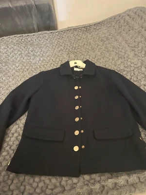 Svart rock från Six Ames med guldknappar - Stilren svart rock från Six Ames med snygga guldknappar framtill och två fickor. Klassisk krage och rak passform, perfekt för dig som gillar en clean look. Jackan är i ullblandning och passar till både höst och vinter.