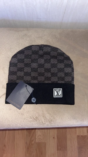 Svart och grå rutig Loui Vuitton mössa - Snygg mössa från Louis Vuitton i svart och grått med klassiskt rutigt mönster och uppvikt kant. LV-logga framtill ger en lyxig touch. Perfekt för dig som vill ha en stilren och trendig accessoar till hösten och vintern.