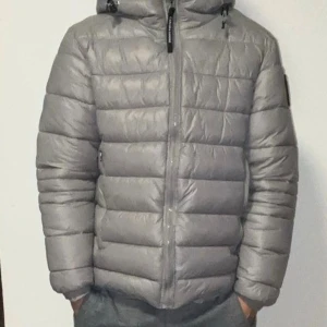 Grå dunjacka Canada Goose - Säljer en grå dunjacka från Canada Goose med huva och reflekterande detaljer. Jackan har ett snyggt quiltat mönster, dragkedja framtill och patch på ärmen. Perfekt för kalla dagar och har en modern, clean look som funkar till streetwear.