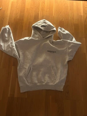 Grå Essentials Fear of God hoodie S - Säljer en grå hoodie från Essentials Fear of God i storlek S. Tröjan har huva, magficka och tryck på bröstet. Perfekt för dig som gillar streetwear och vill ha en clean look. Materialet är mjukt och skönt, troligen bomull och polyester.