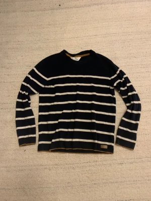 Mörkblå randig tröja från H&M - Snygg mörkblå tröja från H&M med vita horisontella ränder och diskreta bruna detaljer vid hals och nederkant. Tröjan har långärmad design och är tillverkad i mjuk bomull, perfekt för dig som gillar klassisk stil med en twist. Storleken är 152 men passar 160+