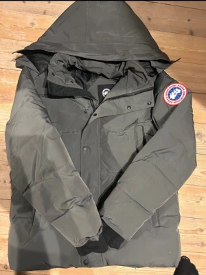 Mörkgrå dunjacka från Canada Goose - Säljer en mörkgrå dunjacka från Canada Goose med ikonisk logga på ärmen. Jackan har huva, dragkedja och knappar framtill samt ribbade muddar vid ärmsluten. Perfekt för kalla vinterdagar och riktigt snygg streetstil.