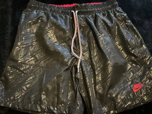 Svarta mönstrade Nike badshorts med snörning - Nike badshorts i svart med glansigt, diskret mönster och röd Nike-logga. Elastisk midja med röd insida och vit/röd snörning. Meshfoder och dragkedja på fickan. 