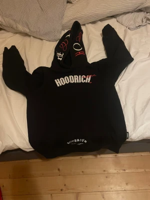 Svart hoodie från Hoodrich med tryck - Svart hoodie från Hoodrich med stor vit logga på bröstet och mindre tryck vid nederkanten. Huvan har coola röda och vita detaljer. Tröjan har lång ärm och klassisk passform, perfekt för dig som gillar streetwear och vill sticka ut. Den är inte använd mycket alls. Kanske tre tvättar!