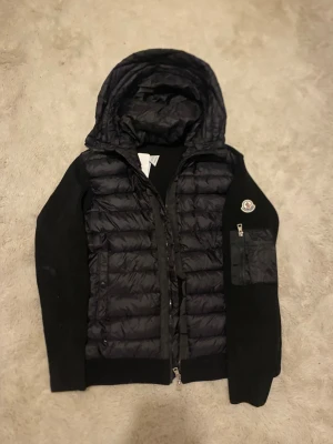 Svart Moncler pufferjacka med huva - Svart pufferjacka från Moncler med stickade ärmar och quiltad front. Jackan har en stor huva, dragkedja framtill och en ficka med dragkedja på ärmen. Moncler-logga på vänster ärm. Perfekt för dig som gillar exklusiva streetwear-vibbar.