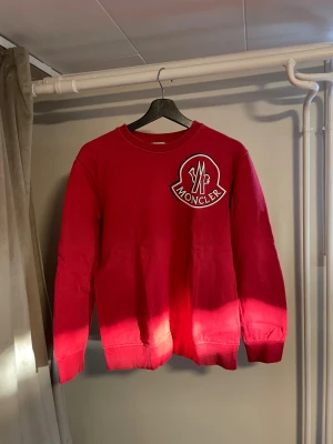 Moncler Sweatshirt – Storlek S - Säljer en röd Moncler sweatshirt i storlek S. Tröjan är i bra begagnat skick, med viss naturlig urtvättning efter användning – vilket syns lite på färgen men påverkar inte passform eller komfort. Stort Moncler-patch på bröstet samt den karakteristiska randiga detaljen längs ryggen.