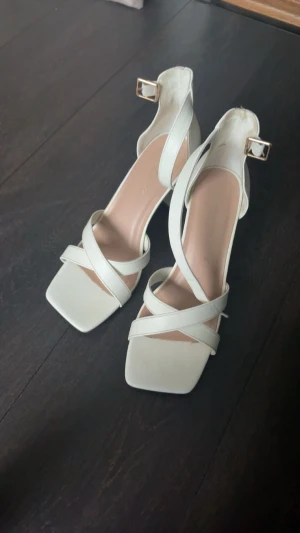 Vita sandaler med klack och remmar - Snygga vita sandaler med bred klack och korsade remmar över foten. Skorna har fyrkantig tå och guldfärgat spänne vid ankeln. Tillverkade i skinnimitation med en stilren och modern look, perfekta för att lyfta din outfit.