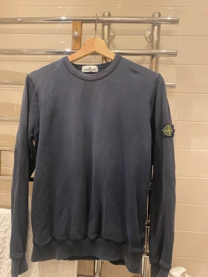 Mörkblå Stone Island sweatshirt - Snygg mörkblå sweatshirt från Stone Island Junior med klassisk rund hals och lång ärm. Tröjan har den ikoniska logotypen på vänster ärm och är gjord i mjuk bomull. Perfekt för dig som gillar streetwear och vill ha en clean look.
