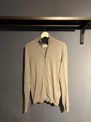 Grå fullzip/cardigan från Pier One - Grå fullzip/cardigan från Pier One med hel dragkedja framtill och ribbad krage. Tröjan har långa ärmar och är tillverkad i mjukt material som känns skönt mot huden. Perfekt för lager-på-lager och chill dagar. Snygg och enkel att matcha med jeans eller joggers.