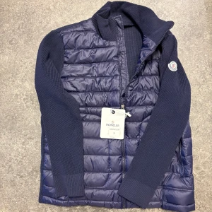  Moncler Cardigan - Säljer en mörkblå pufferjacka från Moncler med stickade ärmar och quiltad framsida. Jackan har dragkedja, ribbad krage och Moncler-logga på ärmen. Insidan har en cool serietidningsetikett. Perfekt för dig som vill ha en snygg och bekväm jacka med street vibe.