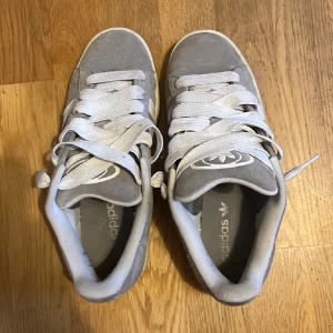 Grå Adidas Campus sneakers mocka - Säljer ett par grå Adidas Campus sneakers i mocka med klassiska vita ränder på sidorna och vit sula med gummidetaljer undertill. Skorna har vit snörning och låg profil, perfekt för en clean och stilren look. Adidas-logga på plösen och insidan.