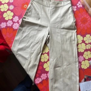 Beiga ”skinn”byxor med dragkedja - Snygga beiga byxor i skinnimitation med raka ben och markerade sömmar framtill. Byxorna har en dragkedja i sidan. Se sista bild för passform.