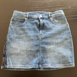 Blå jeanskjol från Gant med logoband - Snygg blå jeanskjol från Gant med klassisk femficksdesign och logoband i mörkblått längs sidorna. Kjolen har bälteshällor, knapp och dragkedja framtill samt Gant-läderpatch bak. Perfekt för en cool och avslappnad stil.