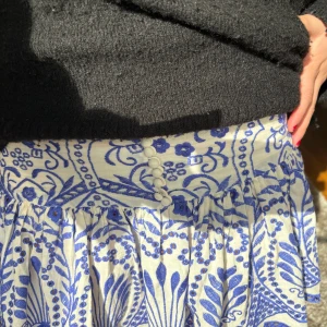 Vit och blå blommig kortkjol med knappar - Supersöt kortkjol i vitt med blått blommigt mönster och dekorativa knappar framtill. Kjolen har en volangkant nedtill och är tillverkad i ett lätt och luftigt material, perfekt för varmare dagar. Sitter snyggt i midjan och har en feminin, flowy siluett💙