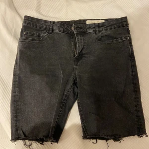 Svarta jeansshorts med rå kant - Svarta jeansshorts från Bershka med längre ben och rå, fransig kant nertill. Klassisk femficksmodell med dragkedja och knapp framtill. Perfekta för en avslappnad och edgy stil. 