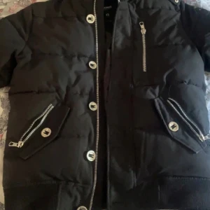 Svart pufferjacka med dragkedjor - Snygg svart pufferjacka med coola silverfärgade dragkedjor och stora knappar. Jackan har flera fickor med dragkedjor och en modern, boxig passform. Perfekt för dig som vill ha en stilren och trendig look under kyliga dagar.