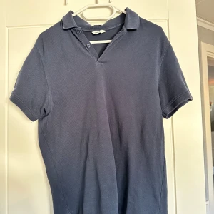 Mörkblå pikétröja från East West - Snygg mörkblå pikétröja från East West i ekologisk bomull. Klassisk krage med två knappar och korta ärmar. Perfekt för en clean och avslappnad stil. Passar dig som gillar enkelhet och kvalitet.