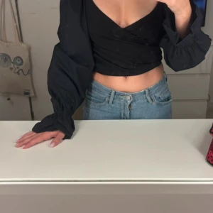 Svart omlottopp med puffärmar H&M - Säljer en svart croppad omlottopp från H&M med långa puffärmar och resår vid ärmslut. Toppen har djup v-ringning och är tillverkad i ett lätt, vävt material. Perfekt för dig som vill ha en trendig och stilren look.