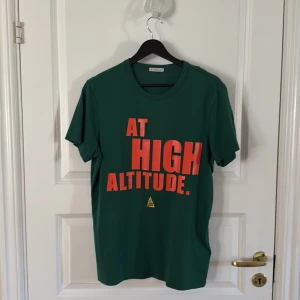 Grön Moncler t-shirt med tryck - Snygg grön t-shirt från Moncler med stort orange tryck 'AT HIGH ALTITUDE.' på bröstet och klassisk Moncler-logga på ärmen. Tillverkad i mjuk bomull och har en normal passform. Perfekt för dig som gillar streetwear och vill sticka ut.
