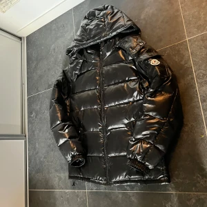 Svart pufferjacka från M Fashion - Snygg svart pufferjacka från M Fashion med glansig finish och huva. Jackan har dragkedja framtill, två fickor med dragkedja och en patch på ärmen. Perfekt för kalla dagar och ger en clean streetstyle-look.