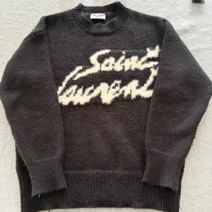 Svart Saint Laurent stickad tröja - Säljer en svart stickad tröja från Saint Laurent med vit logga framtill. Tröjan har rund halsringning, ribbade muddar och är långärmad. Perfekt för dig som vill ha en statement piece med lyxig känsla.