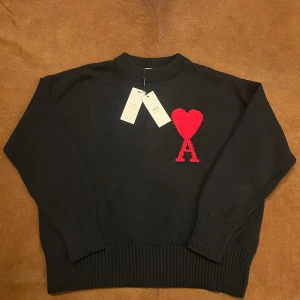 Svart stickad tröja från Ami - Svart stickad tröja från Ami Alexandre Mattiussi med röd hjärtlogga och bokstaven A på bröstet. Tröjan har ribbade muddar och rund hals. Perfekt för dig som gillar stilrena plagg med en twist.