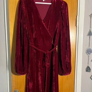 Vinröd sammetsklänning med omlott - Vinröd knälång klänning i mjuk sammet med omlottmodell och v-ringning. Klänningen har långa puffiga ärmar och ett matchande knytbälte i midjan.