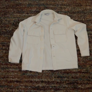 Vit overshirt från Zara - Säljer en stilren vit overshirt från Zara med klassisk krage, stora bröstfickor med lock och knäppning framtill. Jackan har raka ärmar och är perfekt att slänga över en hoodie eller t-shirt för en clean look.