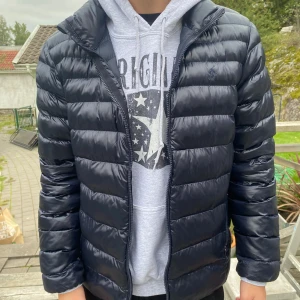 Mörkblå pufferjacka från Polo Ralph Lauren - Snygg mörkblå pufferjacka från Polo Ralph Lauren med horisontella quiltade paneler och dragkedja framtill. Jackan har hög krage, två sidofickor med dragkedja och diskret logga på bröstet. Perfekt för kyliga dagar.