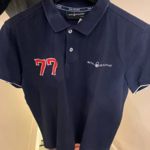 Marinblå Sail Racing piké S - Snygg marinblå pikétröja från Sail Racing med vit logga på bröstet och röd broderad 77. Klassisk krage, korta ärmar med vit kant och knappar framtill. Perfekt för dig som gillar sportig stil och vill sticka ut med coola detaljer.