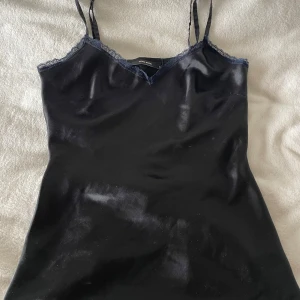 Svart satinlinne med spets från Vero Moda - Snyggt svart linne från Vero Moda i glansigt satinmaterial med tunna justerbara axelband och spetsdetaljer längs v-ringningen. Perfekt att styla med jeans eller kjol för en trendig look.