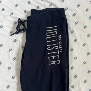 Marinblå vintage mjukisbyxor från Hollister M - Vintage marinblåa mjukisbyxor från Hollister med stort tryck längs ena benet. Byxorna har dragsko i midjan för skön passform och är gjorda i mjukt bomullsmaterial. Perfekta för chill eller träning, med klassisk Hollister-vibe.