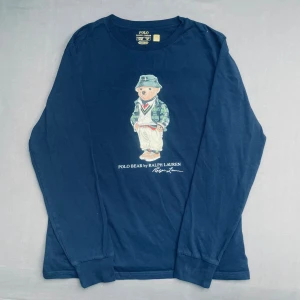 Mörkblå långärmad Polo Bear tröja - Snygg mörkblå långärmad tröja från Ralph Lauren med ikoniska Polo Bear-trycket på bröstet. Rund hals och mjukt bomullsmaterial som känns skönt mot huden. Perfekt för dig som gillar streetstyle och klassiska märken. Storlek XL för barn så passar som ungefär M
