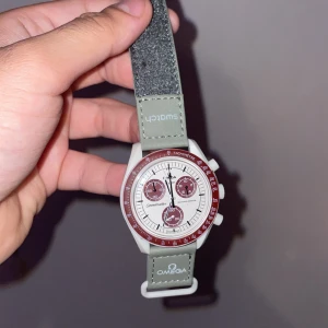 Omega x Swatch Speedmaster MoonSwatch - Säljer en Omega x Swatch Speedmaster MoonSwatch med vit urtavla, vinröda detaljer och grått textilband. Klockan har kronograf och tachymeter, samt tydliga loggor på bandet. En riktigt snygg och unik klocka för dig som gillar cool design.