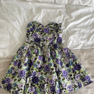 Blommig kort klänning från Divided - Superfin axelbandslös klänning från Divided med blommigt mönster i lila, blått, grönt och svart. Klänningen har en markerad midja med bälte och dragkedja bak. Perfekt snurrig kjol och söt sweetheart-ringning. Materialet känns som bomull.