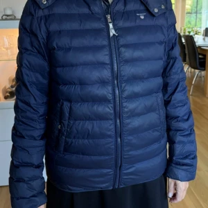 Mörkblå pufferjacka från GANT - Snygg mörkblå pufferjacka från GANT med horisontella quiltade partier och hög krage. Jackan har dragkedja framtill, tryckknappar vid kragen och två sidofickor. Perfekt för kalla dagar och har en stilren look med diskret logga på bröstet.