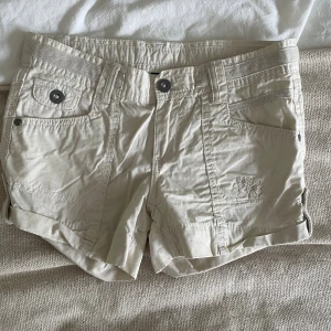 Beige shorts från KappAhl med fickor - Snygga beige shorts från KappAhl med flera fickor fram och bak, knappstängning och bälteshällor. Broderad detalj på ena benet och uppvikta benslut. Perfekta för varma dagar och enkel att matcha med allt.