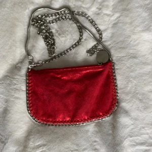 Röd axelväska med kedja - Cool röd axelväska med glansig yta och silverfärgad kedja som axelrem. Väskan har dragkedja upptill och snygga detaljer med silverkedja runt kanterna. Perfekt storlek för det viktigaste och ger en edgy vibe till din outfit.