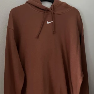 Brun hoodie från Nike med huva - Säljer en brun hoodie från Nike med klassisk vit swoosh-logga på bröstet. Tröjan har huva med snörning och långa ärmar. Mjuk och skön, perfekt för chill dagar. Oversized fit för en avslappnad stil. OANVÄND 