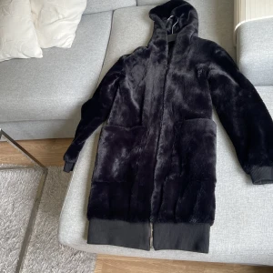 Svart pälsjacka med huva st.M - Säljer en svart pälsjacka med huva och stora fickor framtill. Jackan har lång ärm, ribbade muddar och dragkedja hela vägen. Den är mjuk och fluffig, perfekt för kalla dagar. Längre modell som ger en chill vibe.knappt använd. Mycket bra skick. Hämtas i norsborg botkyrka eller fraktas så fort så möjligt pågrund av flytt.