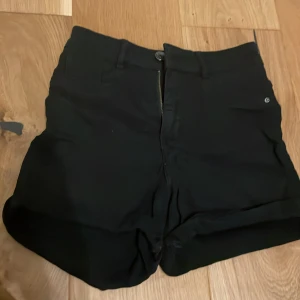 Svarta shorts från Gina Tricot Molly - Snygga svarta shorts från Gina Tricot Molly med klassisk femficksdesign, bälteshällor och dragkedja framtill. Jeanskänsla och uppvikta benslut ger en clean look. Perfekta för varma dagar och enkla att matcha med allt.