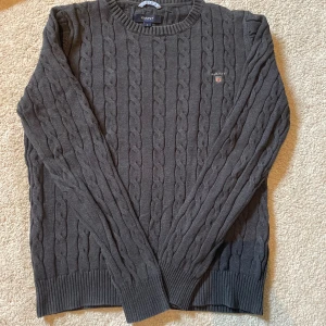 Mörkgrå kabelstickad tröja från GANT - Mörkgrå kabelstickad tröja från GANT med rund halsringning och broderad logga på bröstet. Tröjan har ribbade muddar vid ärmslut och nederkant, och är långärmad. Perfekt för dig som gillar klassisk och stilren look.