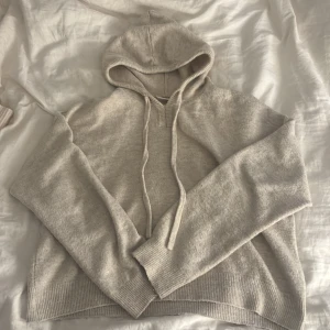 Beige stickad hoodie med huva - Mysig beige stickad hoodie med huva och snörning. Tröjan har långa ärmar och ribbade muddar vid ärmslut och nederkant. Perfekt för kyliga dagar när du vill vara både bekväm och snygg.